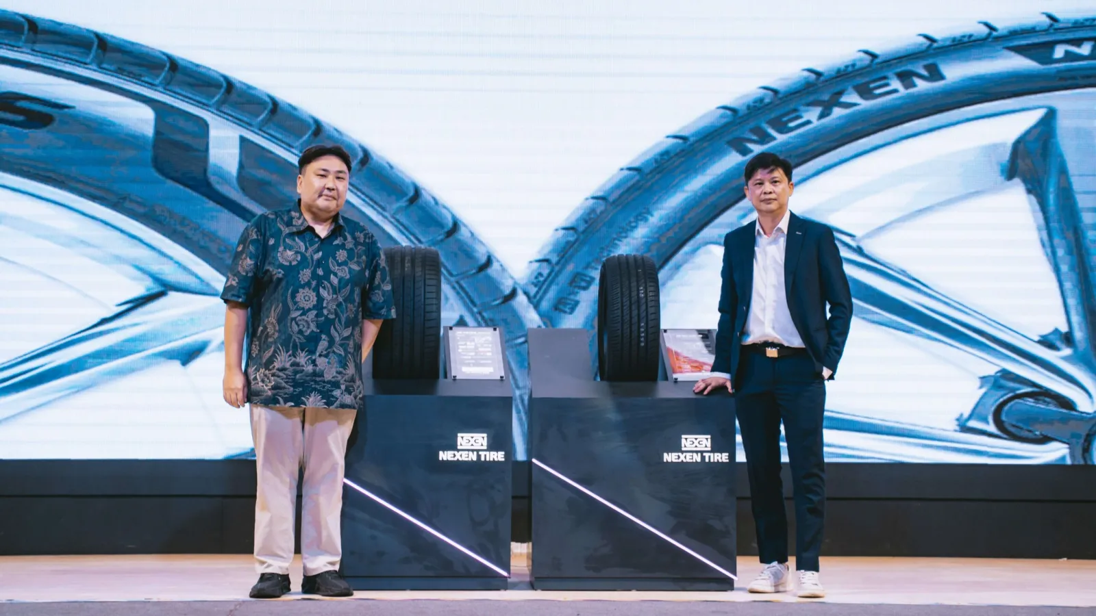 Two male executives posing on stage next to display pedestals holding two black Nexen car tyres in front of a large digital screen backdrop.